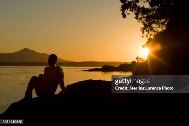 a woman sits on a sandstone bluff in the gulf islands of british columbia to watch a summer sunset over the pacific ocean and the distant mountains of vancouver island near nanaimo, salt spring island, ruxton island, gabriola island and thetis island. - gabriola isle stockfoto's en -beelden