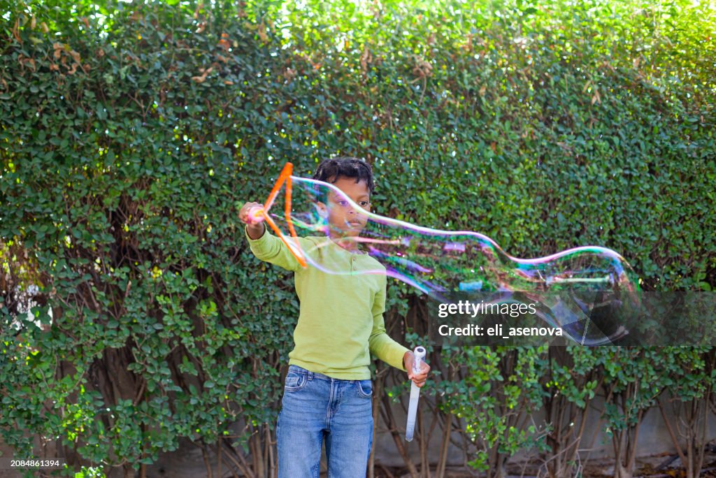 Happy African-American boy with black curly hair blowing and playing with soap bubbles at green garden
