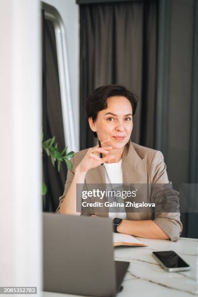 smiling young adult brunette woman in grey suit working at laptop in modern office - brunette sitting at desk stock-fotos und bilder