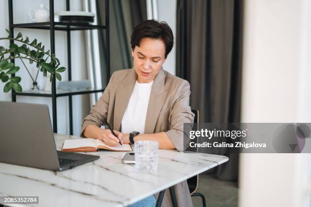 smiling young adult brunette woman in grey suit working at laptop in modern office - brunette sitting at desk stock-fotos und bilder