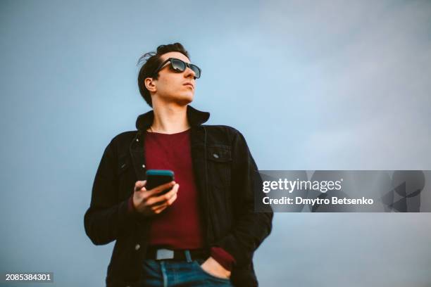 young man looking away while holding smart phone standing against blue sky - real businessman isolated no smile stock pictures, royalty-free photos & images