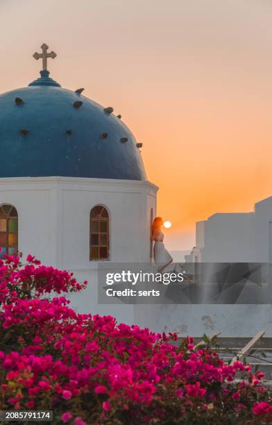 young woman enjoys travelling and looking at the sunset view in the front of a famous traditional blue dome church and red flowers in santorini island, greece thire village, greece - mediterranean-blue-roof-santorini stock pictures, royalty-free photos & images