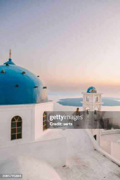 young woman enjoys traveling and looking at the sunset view in the front of a famous traditional blue dome church in santorini island, greece thire village, greece - mediterranean-blue-roof-santorini stock pictures, royalty-free photos & images