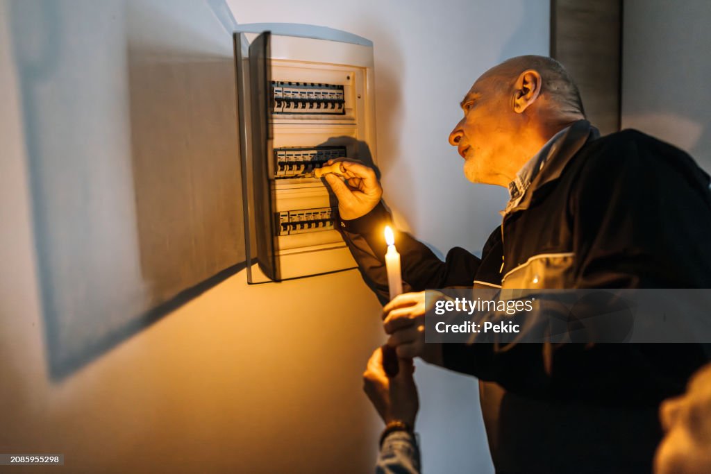 Senior electrician checking fuses for customer at home, customer is holding candlelight