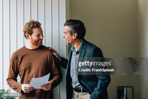 Lächelnder junger Mann und älterer Mann mit Dokumentdiskussion im Büro Lächelnder junger Mann und älterer Mann mit Dokumentdiskussion im Büro