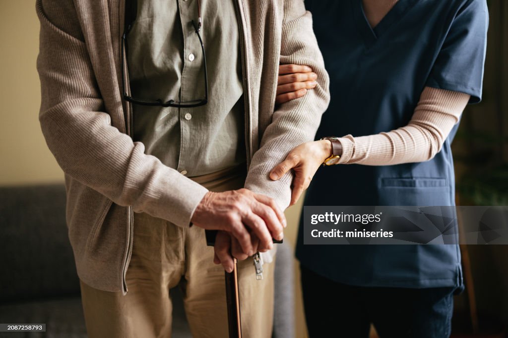 Unrecognizable Nurse Helping Senior Man To Walk In His Living Room