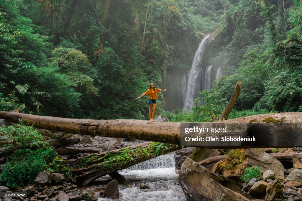 Woman crossing river by log on the background of tropical waterfall while hiking