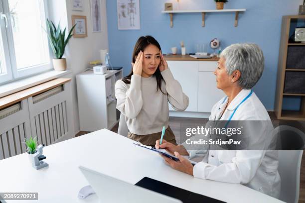 weibliche patientin japanische ethnizität beschreibt kopfschmerzen symptome, zu einer leitenden kaukasischen ärztin - ordination stock-fotos und bilder