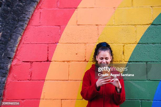 young woman smiling at her phone against a vibrant rainbow mural. - long coat stock pictures, royalty-free photos & images