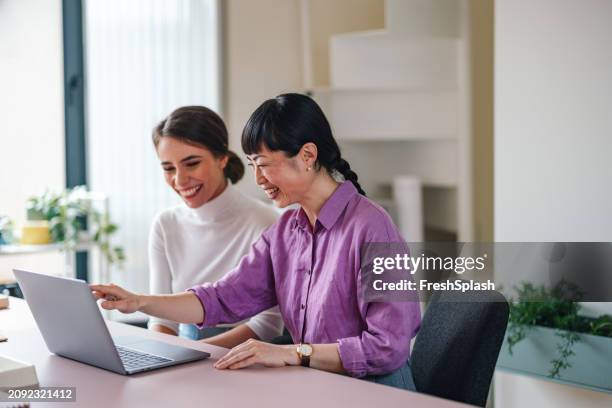 two women laughing and working together on laptop at office - aziatische etniciteit stockfoto's en -beelden