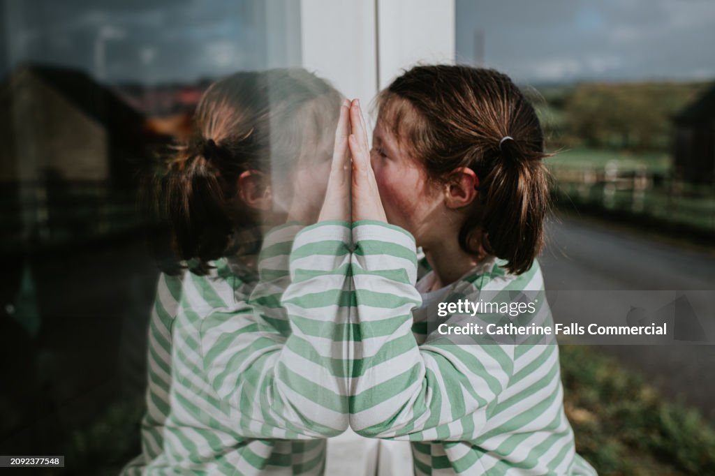 A child pushes herself against a window, nose to nose with a reflection of herself