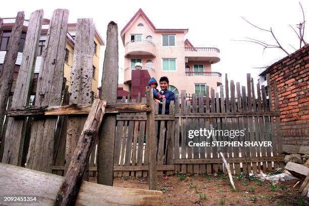 Walking down a street dividing the poor from the wealthy, a father carries his son as they pass the old wooden fence of a typical rural Chinese...