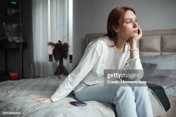 phone anxiety. upset woman sitting on bed with phone looking out window, feeling depressed, waiting for call. social media depression - boredom stock pictures, royalty-free photos & images