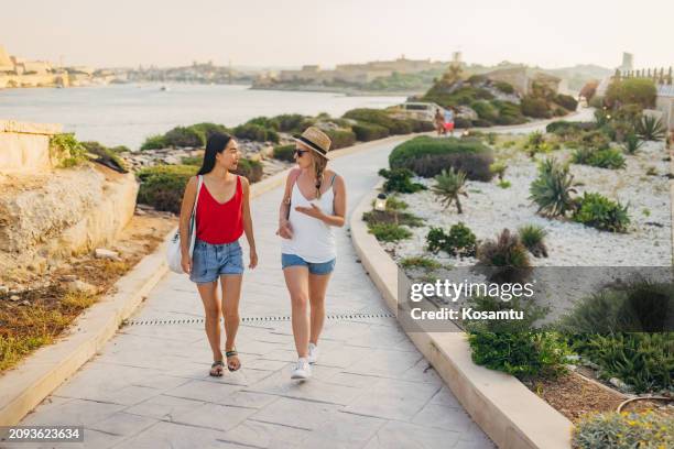 vorderansicht von zwei frauen, die auf einem weg in einem park unweit des meeres und des hafens spazieren gehen - insel malta stock-fotos und bilder
