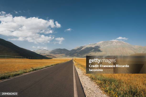 open road through golden fields and mountain range - castelluccio stock pictures, royalty-free photos & images