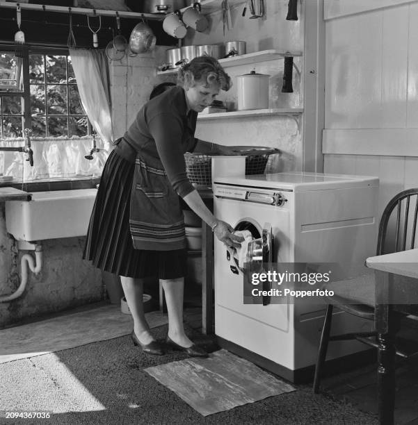 Woman places dirty clothes for cleaning into a front loading automatic washing machine in the kitchen and utility room of her house in England in...