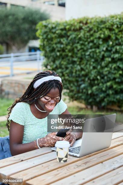 vertical photo of a young black woman student with vitiligo using mobile phone while studying on laptop computer at university campus. concept of youth culture - addiction mobile and laptop stockfoto's en -beelden