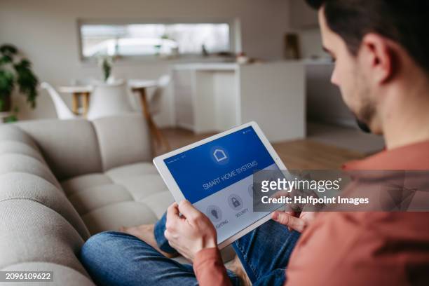 man sitting on sofa, managing features of smart home system. adjusting household functions, lighting, security cameras, door locks, thermostat or heating settings. holding tablet, smart thermostat, rear view. - security cameras stock pictures, royalty-free photos & images