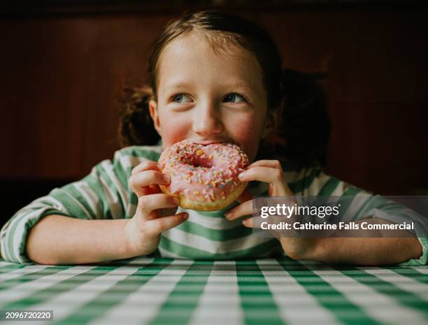 a child smiles as she takes a bite of a pink glazed donut with sprinkles - unhealthy eating stock pictures, royalty-free photos & images