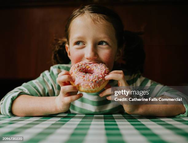 a child smiles as she takes a bite of a pink glazed donut with sprinkles - sticky bun stock pictures, royalty-free photos & images