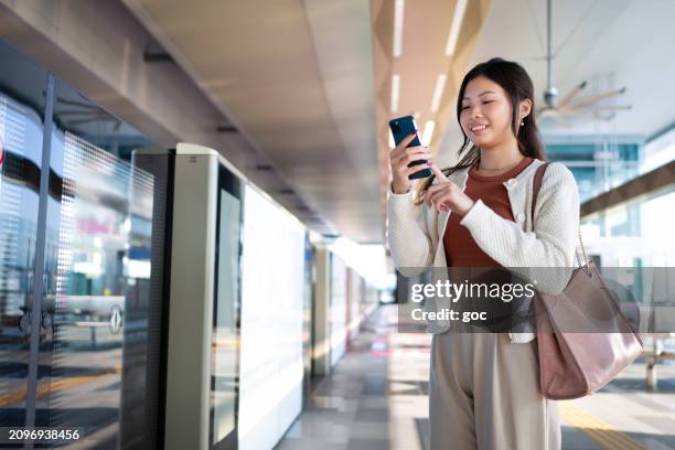 young asian woman using smart phone while waiting for mass rapid transit (mrt) on railway platform - mrt train stock pictures, royalty-free photos & images