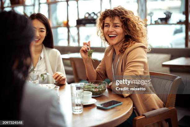 lächelnde, gut gekleidete geschäftsfrau, die lacht, während sie mit kollegen im restaurant zu mittag isst - mittagspause stock-fotos und bilder