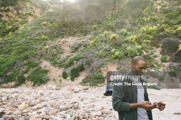 young man using smart phone while standing on rocky beach - malibu nature stock pictures, royalty-free photos & images