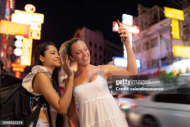 two multiracial female friends enjoying a night walk on the streets of bangkok, thailand, having video call and smiling - friends laughing at iphone video stock pictures, royalty-free photos & images