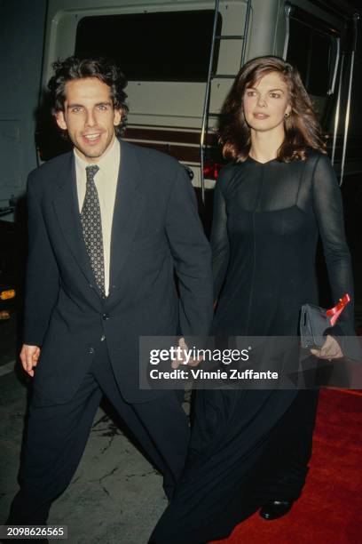 American actor and comedian Ben Stiller, wearing a black suit with a white shirt and patterned tie, holds hands with American actress Jeanne...