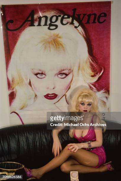 Polish-born American singer, actress and billboard model Angelyne, wearing a low-cut pink minidress with a studded black belt, sits on a black sofa...