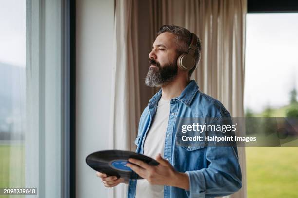 handsome man listening to vinyl with headphones, standing by window. pairing turntable with pair of wireless headphones at home. - stereoanlage stock-fotos und bilder