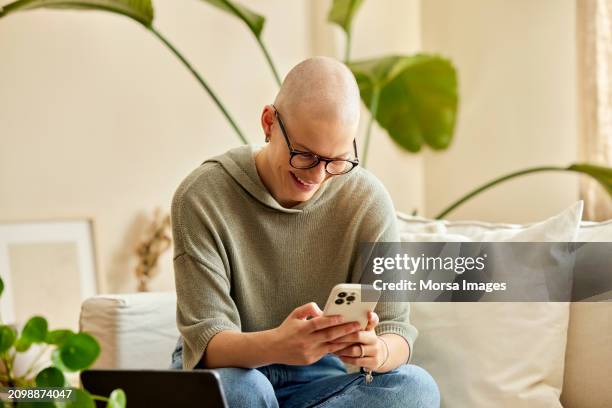 happy female cancer patient using smart phone at home - alopecia stockfoto's en -beelden