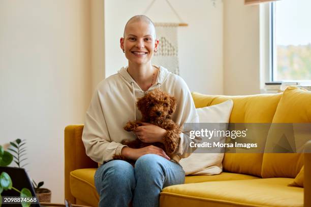 smiling female cancer patient with pet dog on sofa at home - alopecia stockfoto's en -beelden