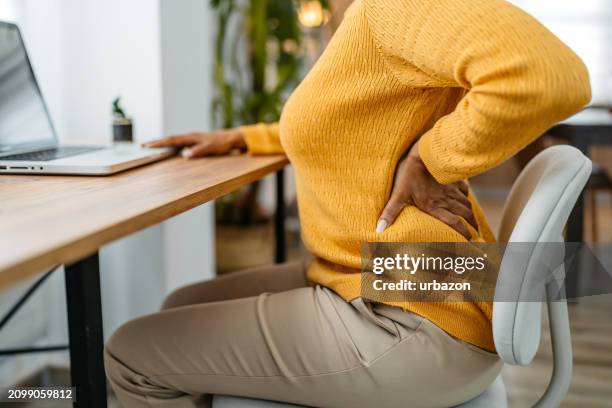 tired young woman having back pain while working on the laptop at home - dolor de espalda fotografías e imágenes de stock