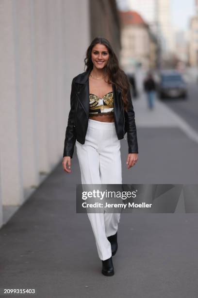 Michelle Golke seen wearing gold necklace, black / white / yellow pattern crop top, black leather biker jacket, white wide leg pants and black...