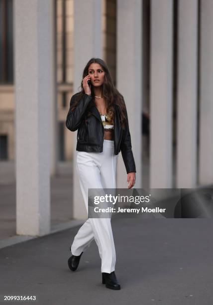 Michelle Golke seen wearing gold necklace, black / white / yellow pattern crop top, black leather biker jacket, white wide leg pants and black...