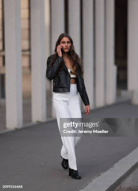 Michelle Golke seen wearing gold necklace, black / white / yellow pattern crop top, black leather biker jacket, white wide leg pants and black...