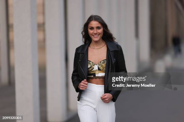 Michelle Golke seen wearing gold necklace, black / white / yellow pattern crop top, black leather biker jacket and white fabric wide leg pants, on...