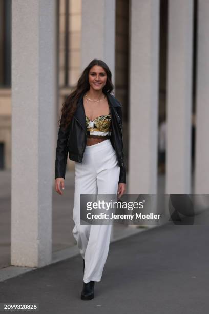 Michelle Golke seen wearing gold necklace, black / white / yellow pattern crop top, black leather biker jacket, white fabric wide leg pants and black...
