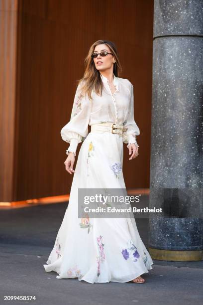 Guest wears sunglasses, a flared and vaporous white long floral dress with a mandarin collar and bishop sleeves, a white double buckle belt, outside...