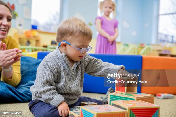 adorable niño pequeño con síndrome de down apila bloques en el aula de preescolar durante una cita de terapia ocupacional - síndrome de down fotografías e imágenes de stock