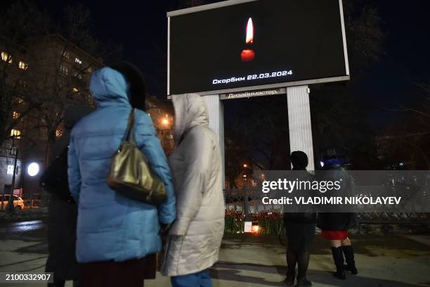 People stand next to an advertising screen displaying an image of a lit candle and the slogan " Mourn " in Novosibirsk on March 23 a day after a gun...