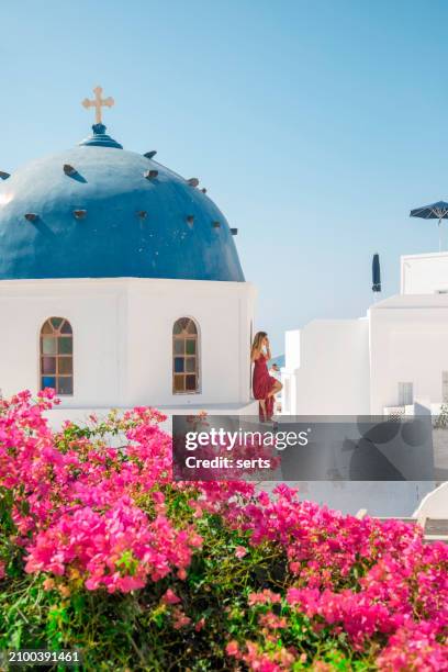 young woman enjoys traveling and looking at the view in the front of a famous traditional blue dome church and red flowers on a sunny summer day in santorini island, greece thira village, greece - mediterranean-blue-roof-santorini stock pictures, royalty-free photos & images