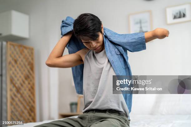 asian young amputee without arm wearing a t-shirt while sitting on bed. attractive strong handicapped man getting clothes with happiness in the morning, enjoying routine preparation in bedroom a home. - young man getting dressed stock pictures, royalty-free photos & images