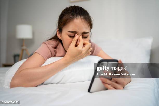 exhausted asian young woman hurt eye while using mobile phone on bed. attractive girl lying down on bed, feeling visual fatigue and eye strain tired from overwork and massage dry irritable eye at home - secco foto e immagini stock