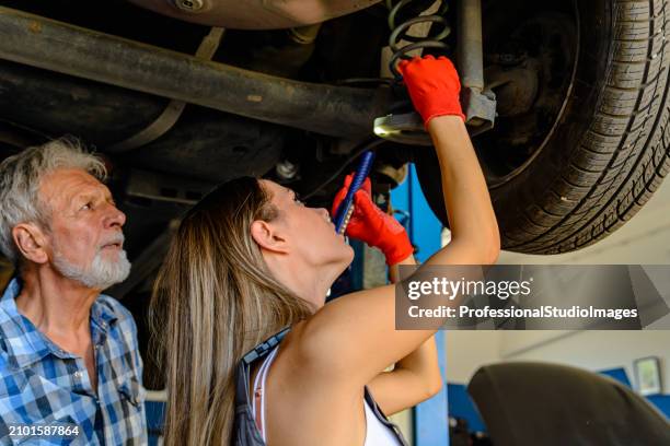 an older car mechanic man and young woman are working underside a car using a mechanic equipment. - car underside stock pictures, royalty-free photos & images