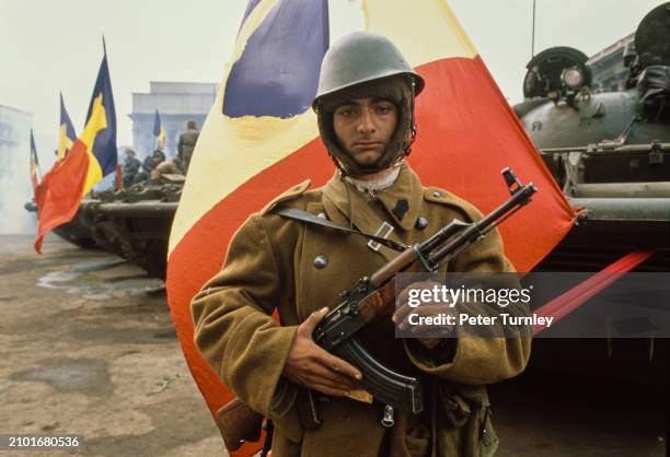 Soldier standing in front of a row of tanks decorated with Romanian flags, Bucharest, December 1989. The flag directly behind him has had the...