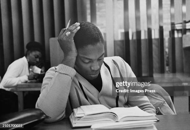 Student, his head resting against his right hand, a pencil held in his fingers, as he reads from a book on the table before him, another student in...