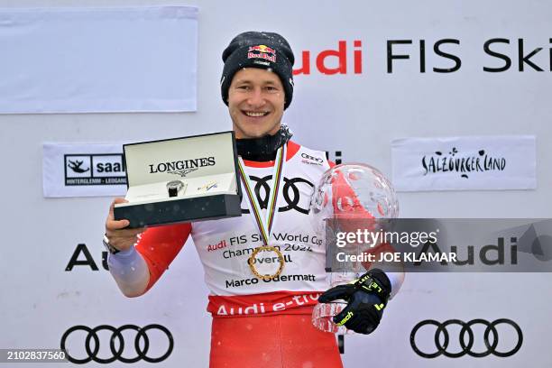 The overall season winner of the Men's FIS Alpine Skiing World Cup Switzerland's Marco Odermatt poses on the podium with his trophy and a watch he...