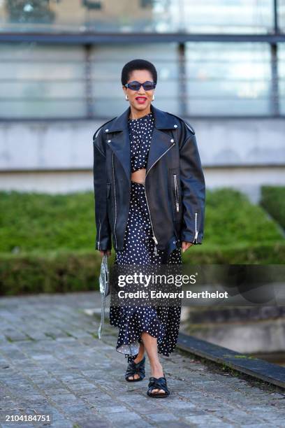 Tamu McPherson wears sunglasses, earrings, a black asymmetric flowing dress with white polka dots and a draped crop top, a black leather biker...